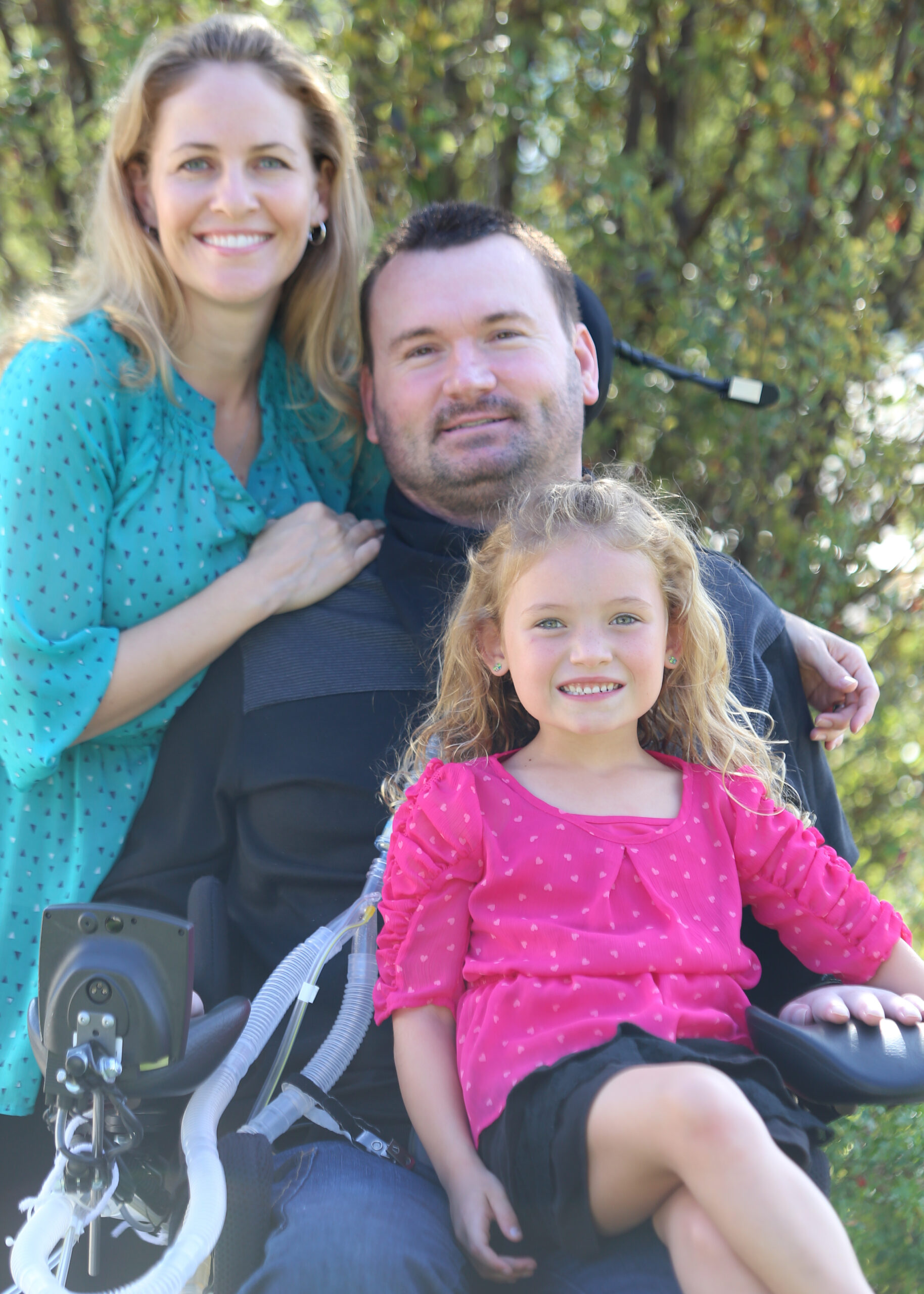Woman with arm around a quadriplegic man with power wheelchair with their young daughter on his lap