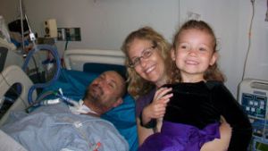 quadriplegic dad is laying in a hospital bed with his wife and daughter next to him