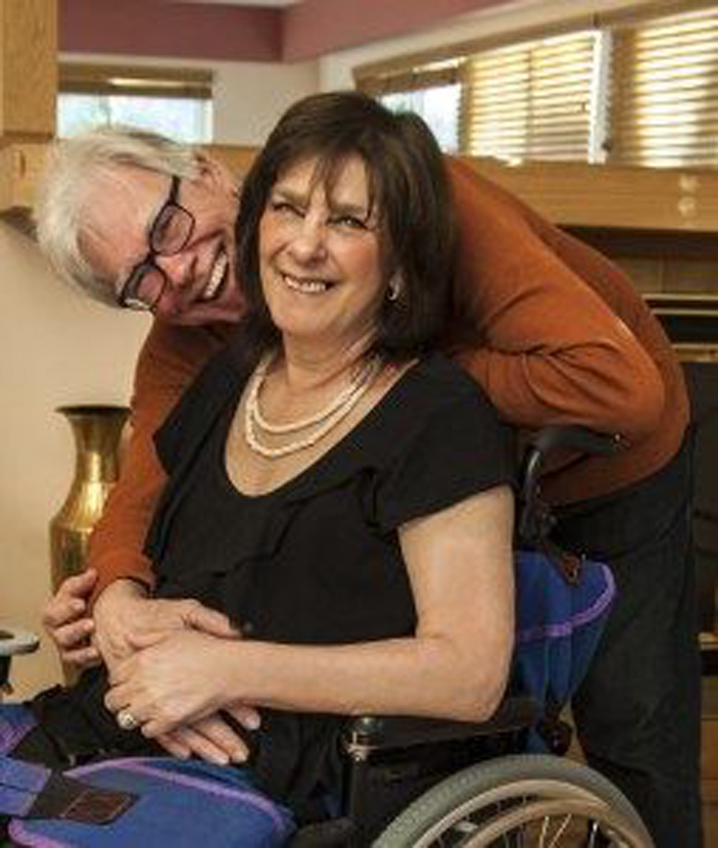 A woman in a wheelchair smiling with her husband hugging her and smiling