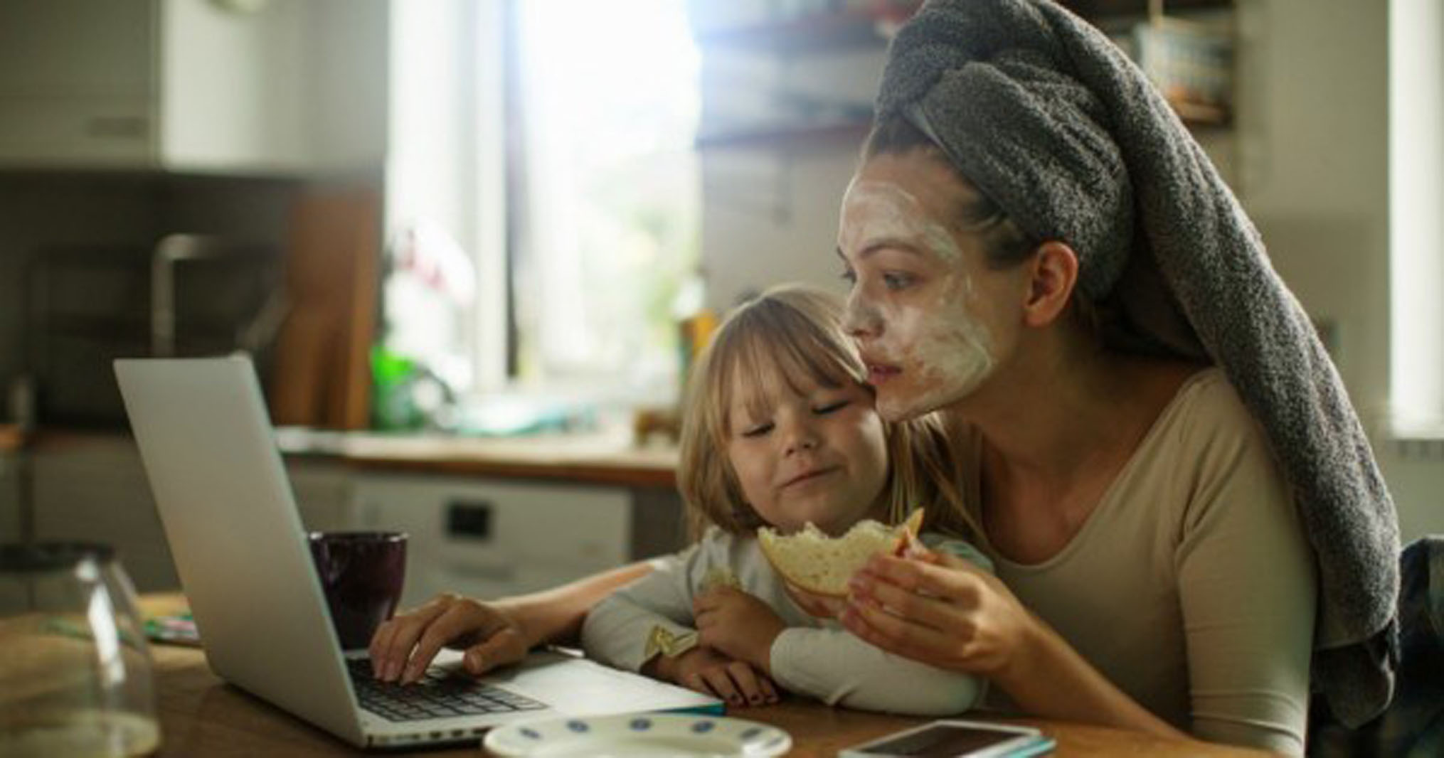 a mom sitting at a table looking at a computer and eating with her daughter on her lap