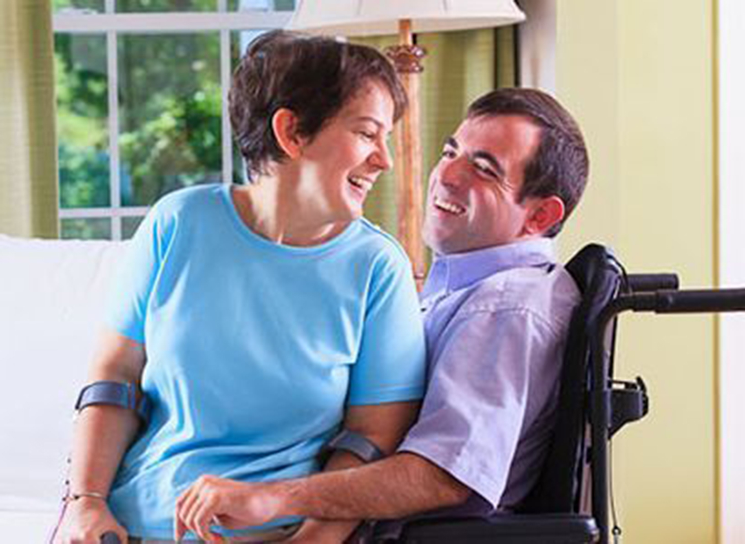 a man in a wheelchair with a woman using crutches on his lap, both smiling and looking at each other