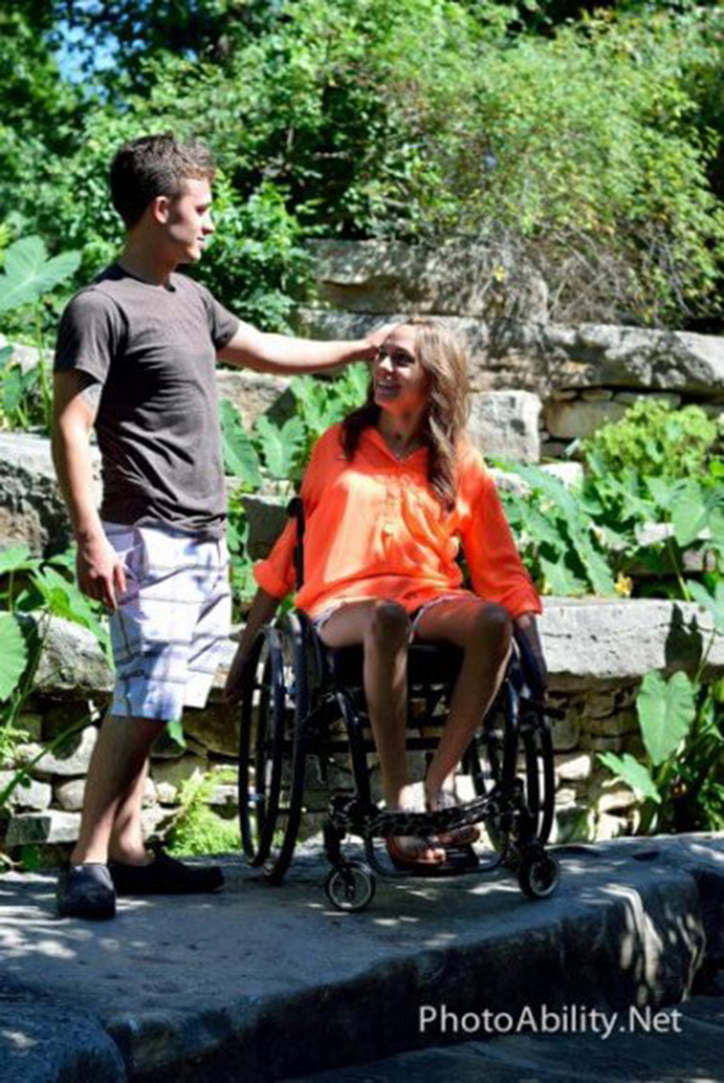 A man standing next to his girlfriend in a wheelchair, them looking at each other, him touching her hair