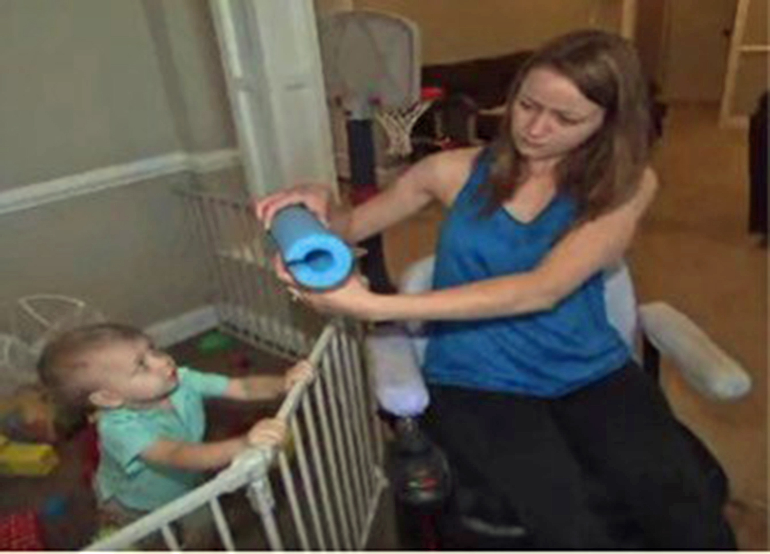 Paraplegic mom holding a pool noodle next to her baby standing in a crib