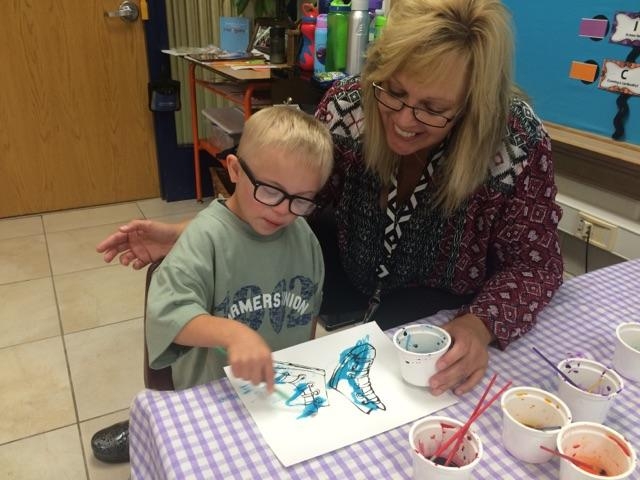 a boy with Down Syndrome paints a picture in class with his teacher