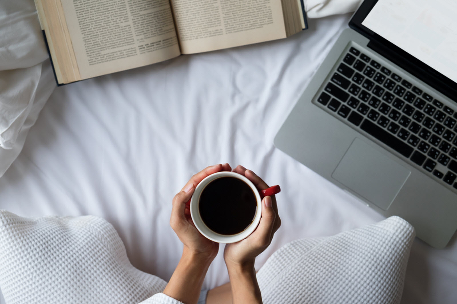 a person sits on a bed holding a cup of coffee next to a laptop and book