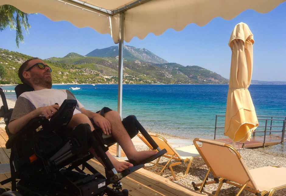 man in power wheelchair tilting back under a tent on an accessible beach with turquoise water
