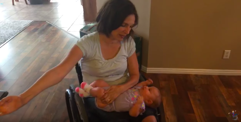 screen shot of meg using wall to propel her wheelchair with one arm on baby in her lap