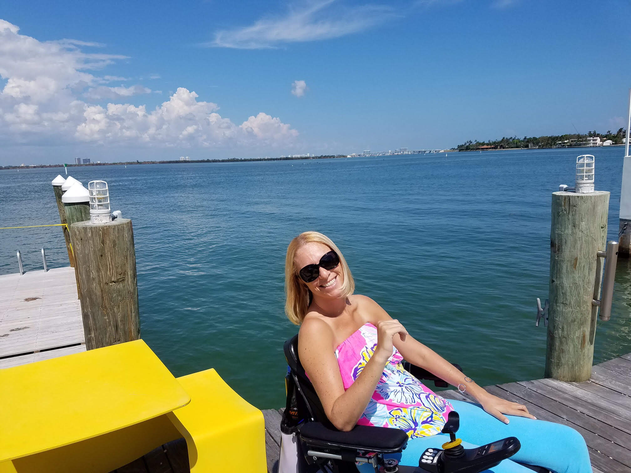Picture of Ali sitting on a dock in her wheelchair, a body of water in the background. She is smiling and has sunglasses on.