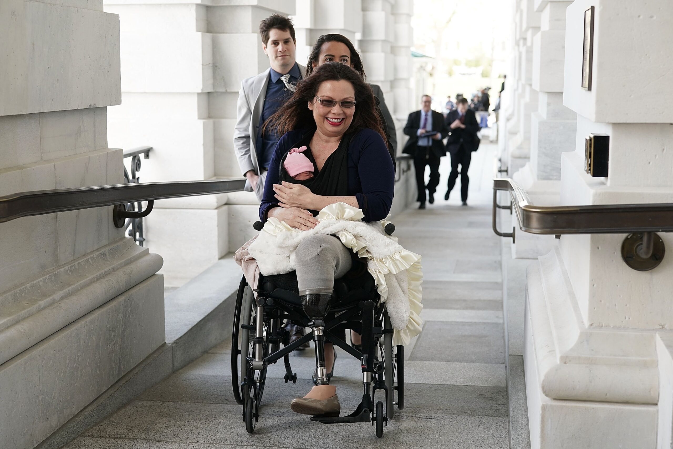 Picture of Tammy Duckworth smiling holding her baby while she sits in her wheelchair.