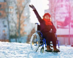Waving happy young adult woman on wheelchair in the city in winter at sunset