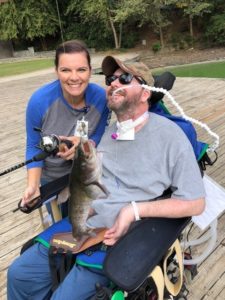 Man in wheelchair showing oiff fish he caught with female companion standing next to him.