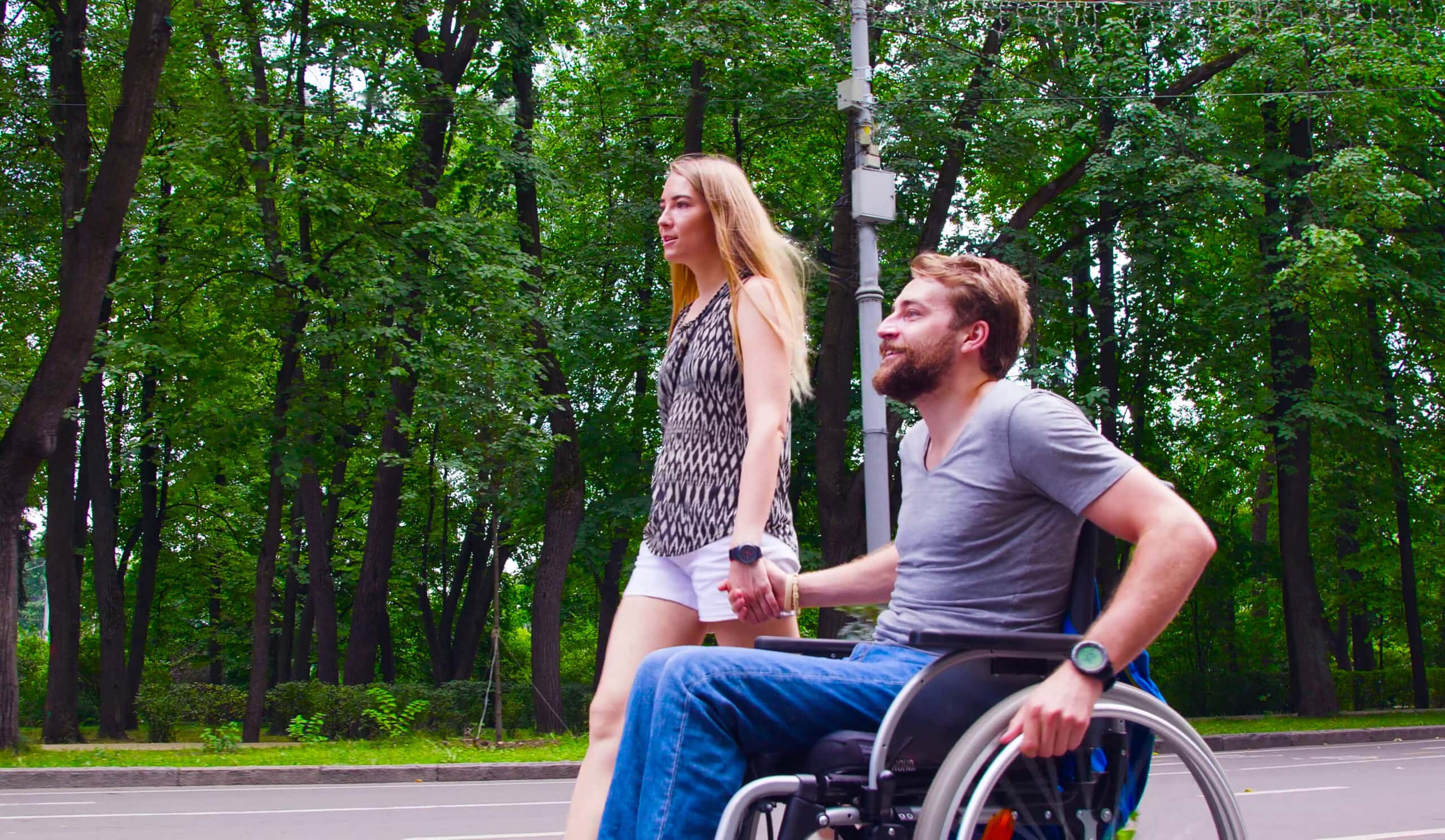Happy young disable man in wheelchair walking in the park with his wife. Side view.