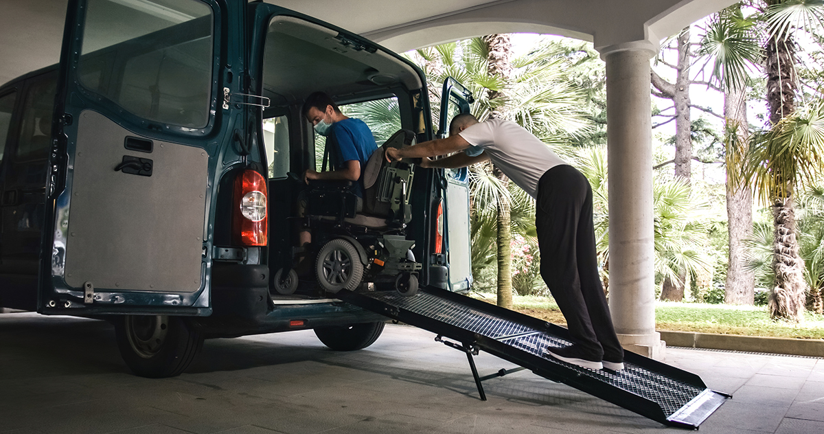 Man on wheelchair using accessible vehicle with ramp for transportation with driver helping.