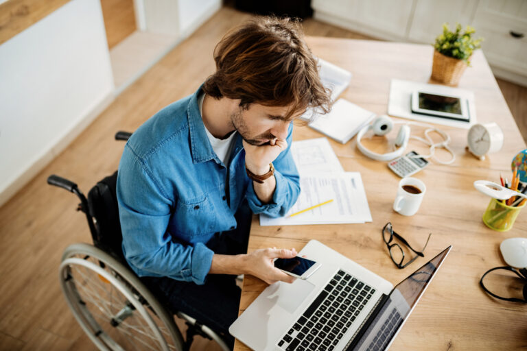High angle view of young entrepreneur in wheelchair texting on smart phone while working on a computer at home.