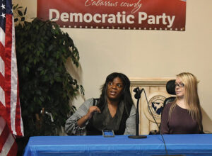 two women in wheelchairs at table behind a microphone next to American flag and Democratic party sign in background