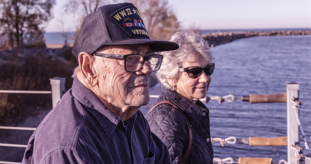 Close-up of a proud, elderly, 93 year old senior adult man USA WWII and Korean Conflict military war veteran and his senior adult woman home caregiver daughter. They are standing side by side sightseeing on a sunny day at a public park on the shoreline of Lake Ontario. He is wearing a common, unbranded, generic souvenir shop military veteran commemorative baseball style cap with generic wording, replica plastic insignia pins and replica campaign ribbon iron-on patches. Photo taken near Rochester, New York, in the Finger Lakes region of western NY State, USA.