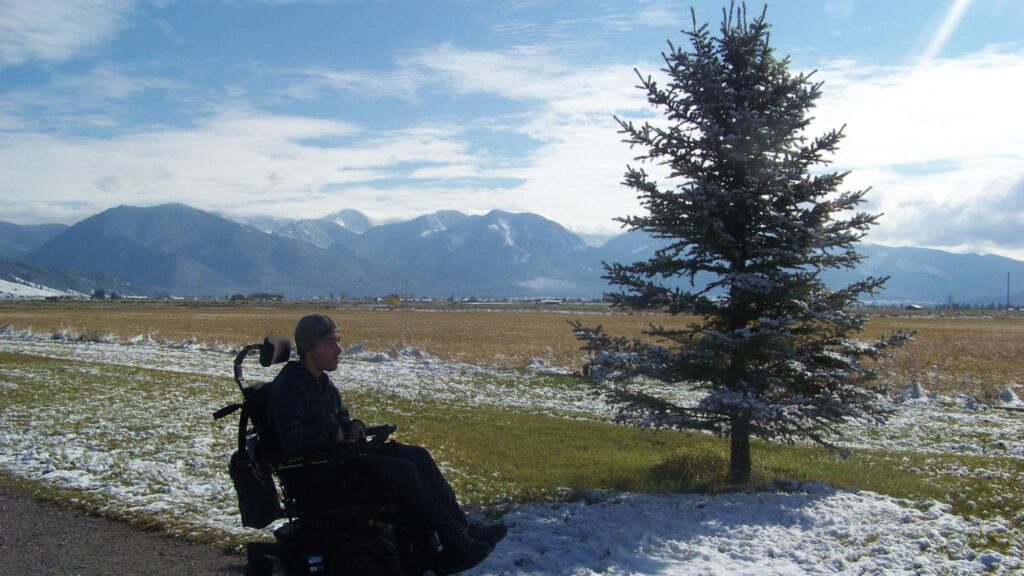 David in powerchair outdoors on the road next to pine tree with mountain range in background 
