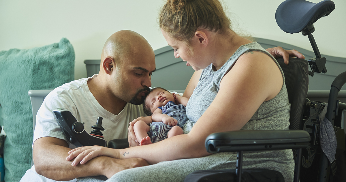 Disabled mother sitting in her wheelchair in front of newborn baby's crib holding her newborn son, husband is kneeling beside her.
