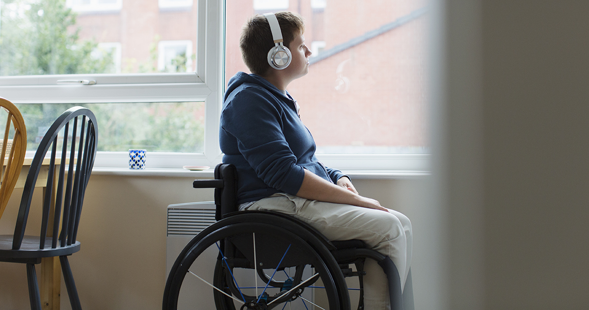 Thoughtful young woman in wheelchair listening to music with headphones at window