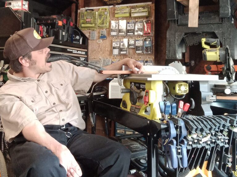 Ouellette in work room using a mouth tool to operate table saw 