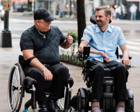 two men in powerchairs outdoors talking. One giving thumbs up