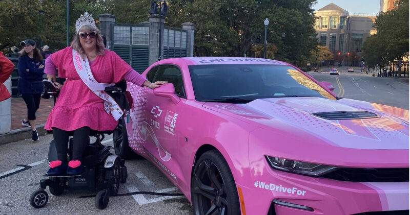 woman in powerchair wearing sash and crown next to a hot pink sports car