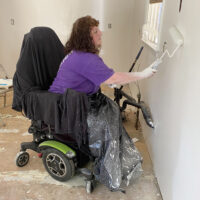 wmoan in powerchair painting the wall of a house. chair and woman covered with tarp