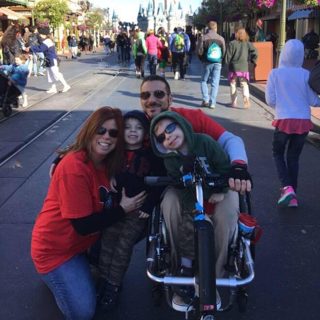 A wheelchair-using dad with his wife and young sons pose for a photo with the Disney castle in the background. 
