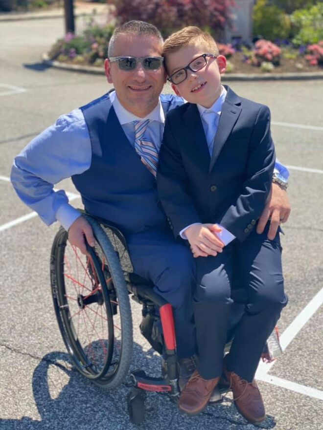 A wheelchair-using dad poses with his young son. Both are wearing suits. 