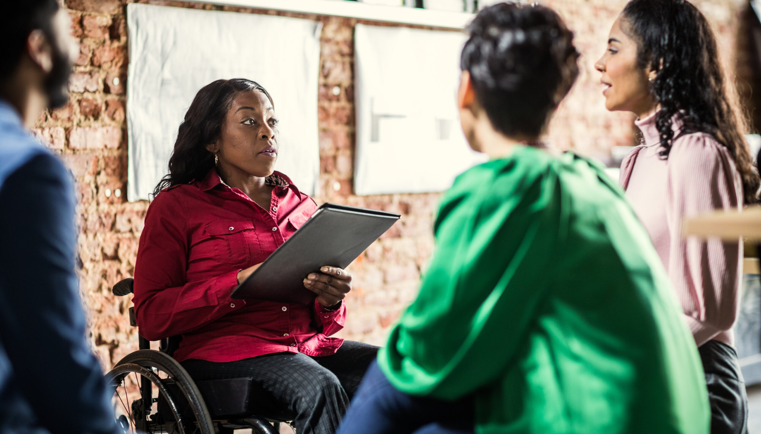 A confident Black woman wheelchair user with a clipboard leads a meeting.