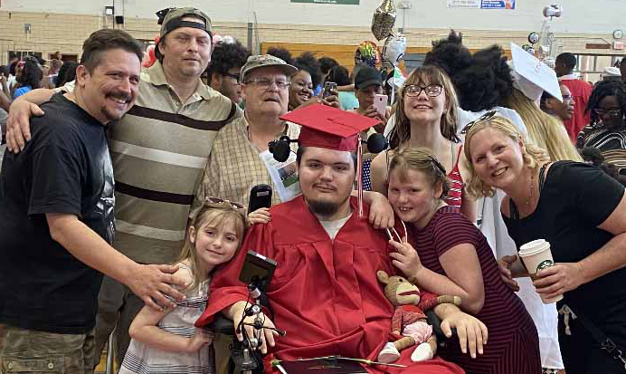 Cyrus in powerchair wearing red cap and gown at his graduation ceremony surrounded by many friends and family