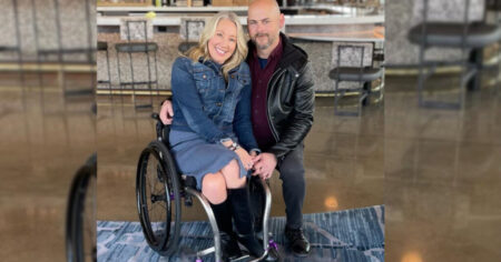 A couple smile at the camera. The woman is a wheelchair user, and the man kneels to be at eye level to her.