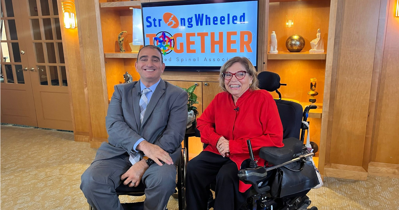 United Spinal Association CEO and Judy Heumann pose in front of a #StrongWheeled Together banner.