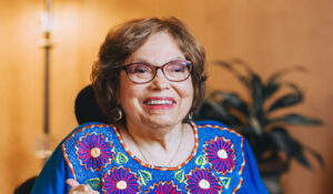 Judy Heumann, a White older woman with glasses, wears a bright blue shirt with purple flowers.