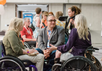 Modeling inclusion, a man wearing glasses and using a wheelchair listens to a woman wheelchair user.