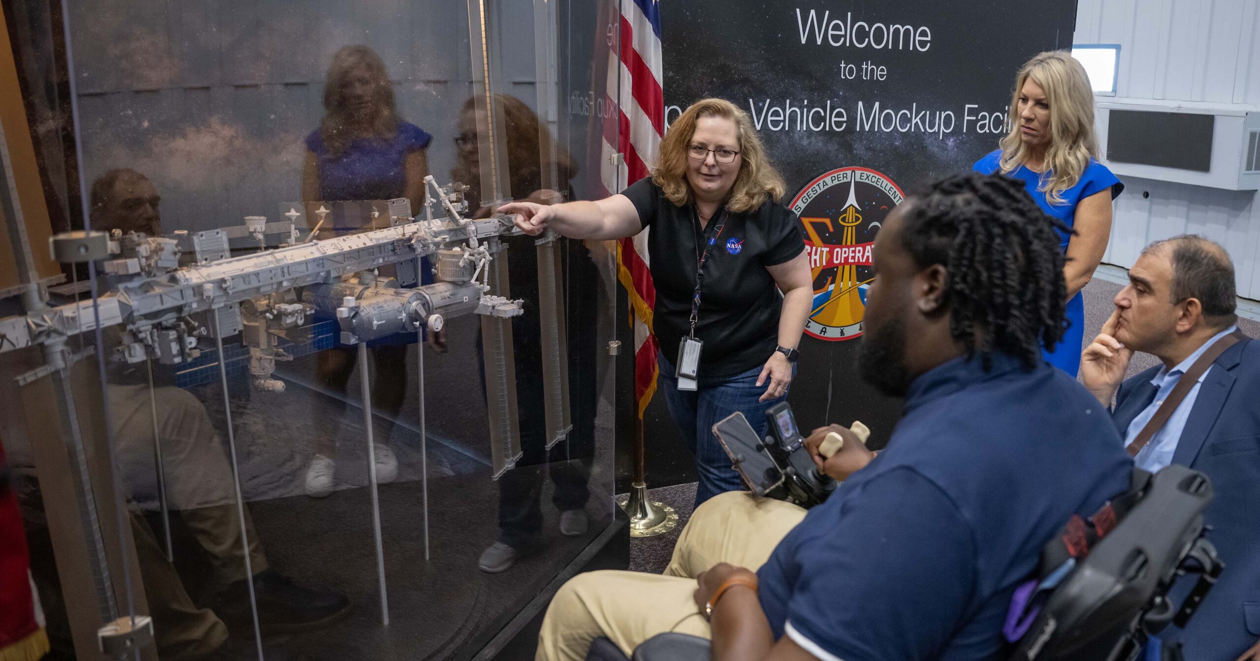 A white woman points out inclusive features on a model of a spacecraft to two men using wheelchairs. One man is Black, and the other is Hispanic.