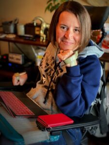A White woman wheelchair user in a blue jacket smiles.