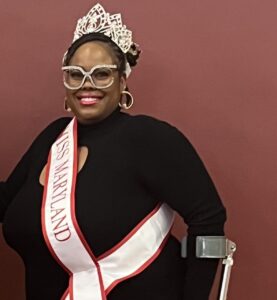 An attractive Black woman wears a crown and a sash, and stands using forearm crutches.
