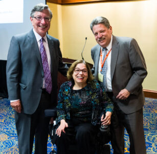 Two white men in suits flank a white woman dressed formally and using a power chair.