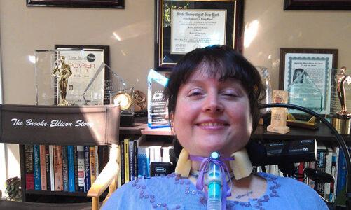 A smiling white woman who uses a vent poses in front of her degrees and awards.