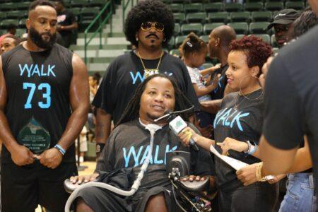 A Black wheelchair user speaks to a reporter. His T-shirt says "walk," and he is surrounded by supporters wearing the same T-shirt.