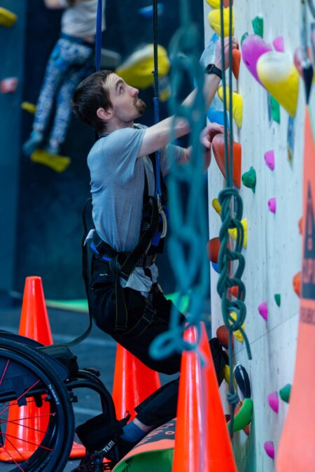 A white man climbs an adaptive rock wall. His manual wheelchair is just below his feet.