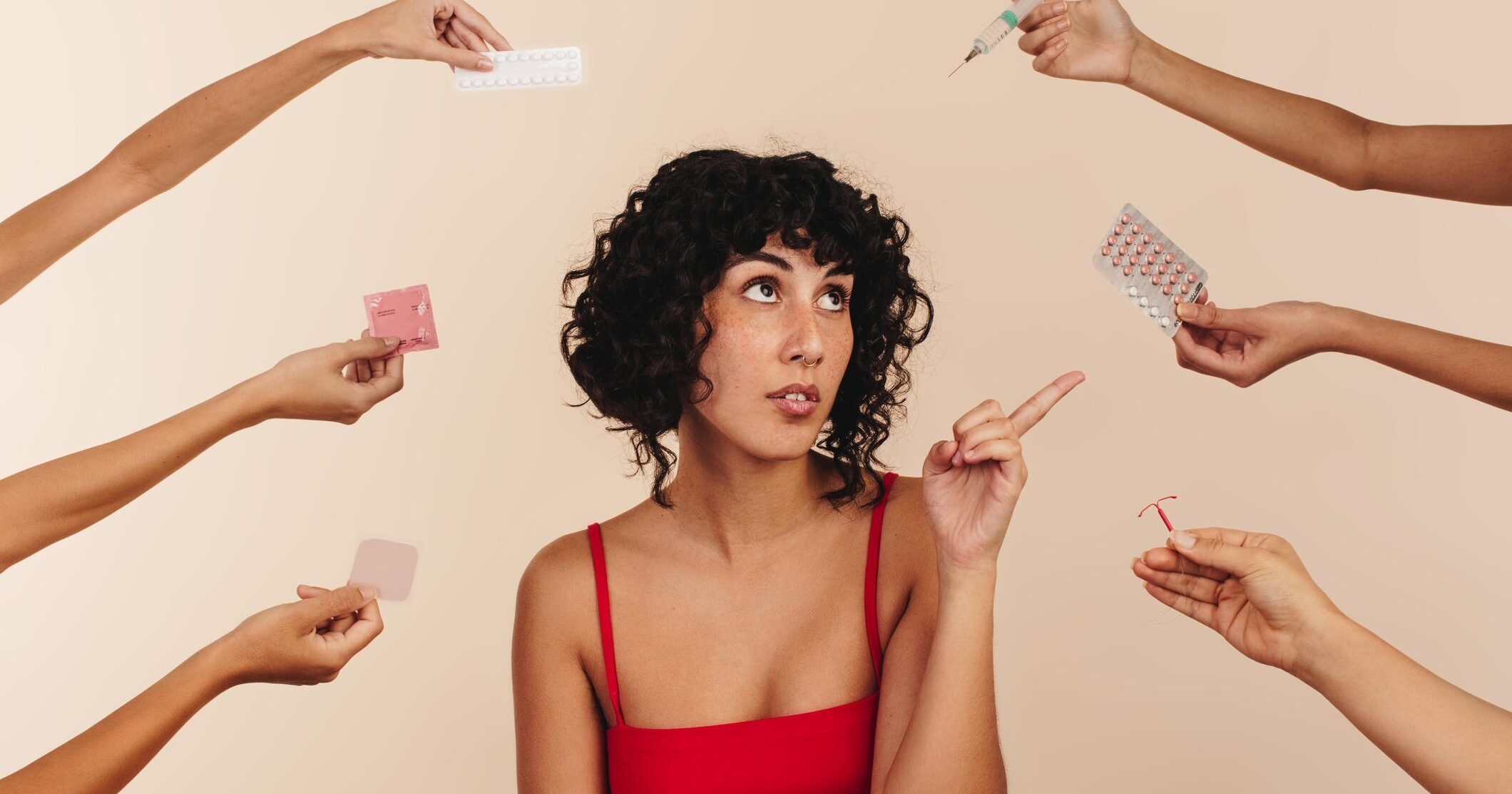 Young woman pointing at a pack of pills while surrounded by hands holding different forms of hormonal and non-hormonal contraception. Modern young woman making choices about her reproductive health.