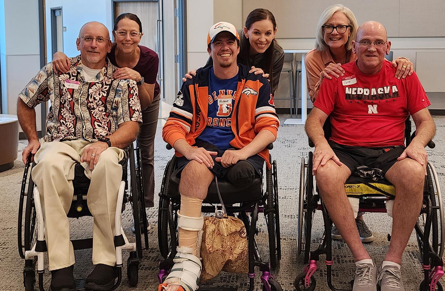 Three men who use wheelchairs smile as three women stand behind them.