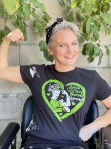 A white woman wheelchair user flexes her arm like Rosie the Riveter. Her T-shirt says, "Spinal Cord Injury Warrior -- Unbreakable." 