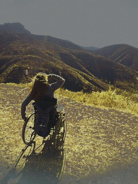 A photo of Laurie Crosby in her wheelchair. The photo is taken behind her and she is looking at mountains with her hand blocking her face from the sun.