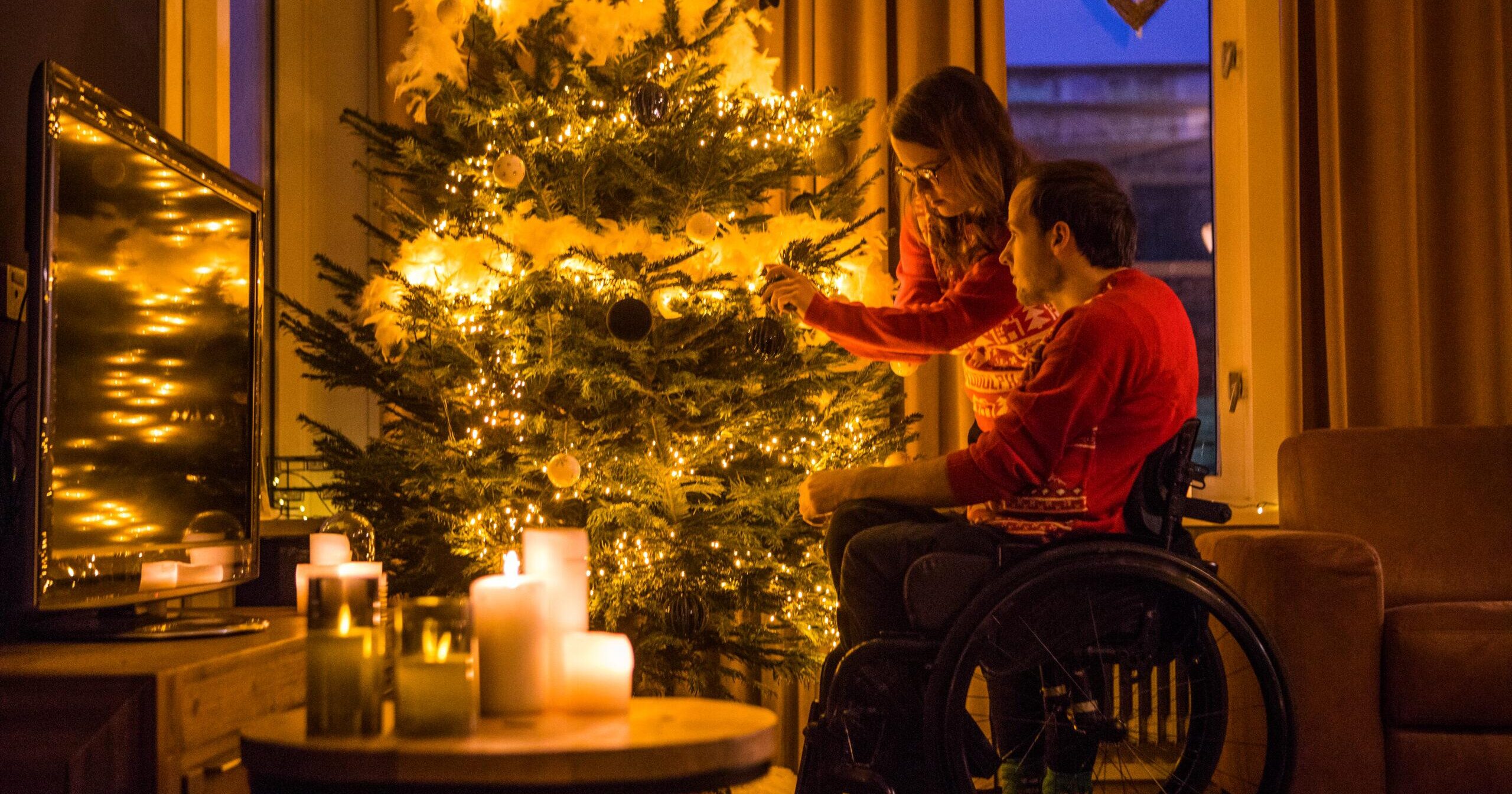 A man sits in a wheelchair with a woman standing next to him by a christmas tree