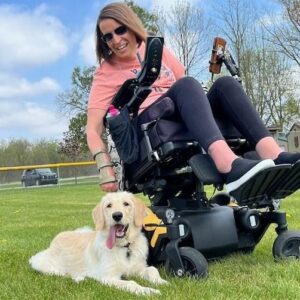 Annie Streit, a white woman in a power wheelchair sits in her power wheelchair at a park with her dog