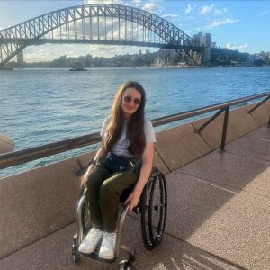 Shannon Kelly, a white woman in a manual wheelchair sits in front of the Sydney Opera House.