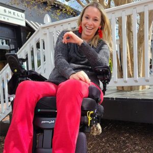 Ali Ingersoll a white woman with blonde hair sits in her power wheelchair in front of her house.
